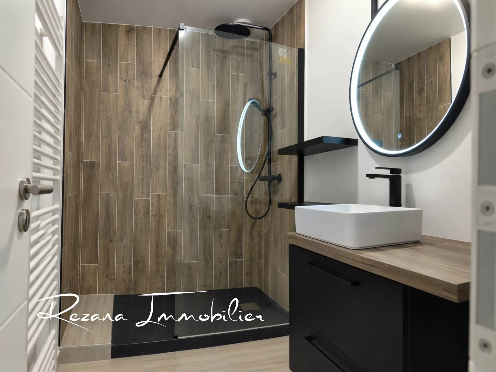 Salle d'eau avec douche, robinetterie noire, carrelage imitation bois et vasque blanche.